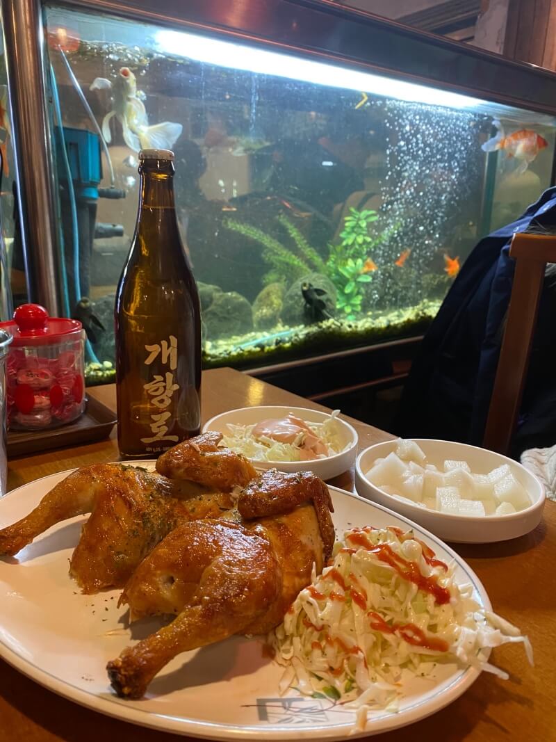통닭 한마리와 인천 개항로 맥주가 놓여있고 뒤에는 물고기들이 있는 어항이 보임